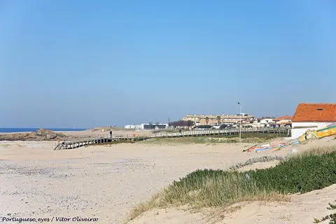 Praia do Marreco com areal, passadiço de madeira, dunas com vegetação e casas ao fundo
