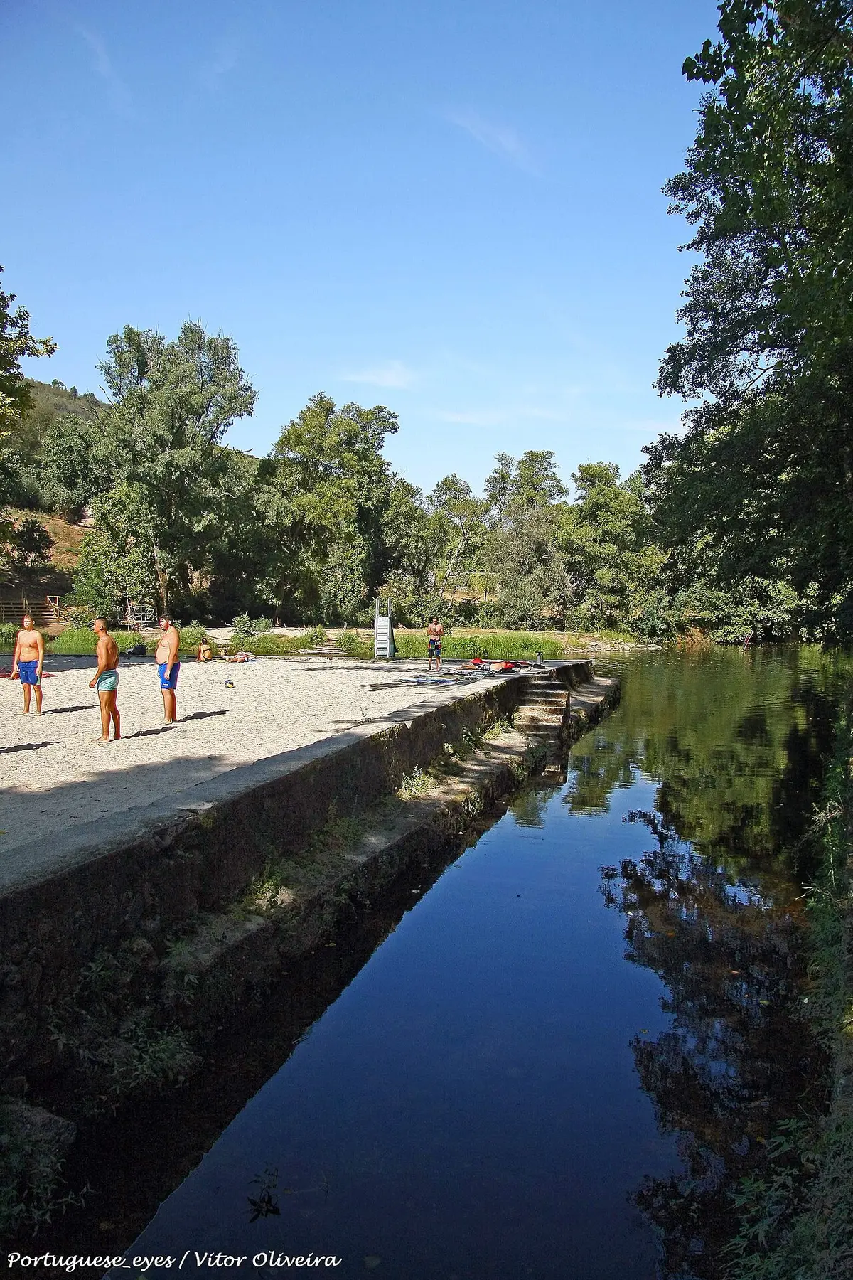 Praia fluvial de São Gião com areal, muro de pedra e rio ladeado por árvores.