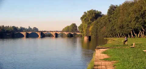 Rio calmo junto a margem relvada arborizada, com ponte de arcos de pedra ao fundo