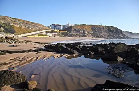 Praia de São Bernardino com rochas, poça de maré na areia e casario no alto da arriba.