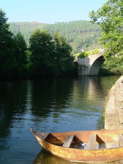 Rio com barco de madeira junto à margem e ponte de arcos em pedra ao fundo, rodeada de vegetação