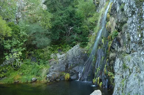 Cascata do Poço do Inferno a cair sobre rochas com vegetação, formando uma poça de água escura