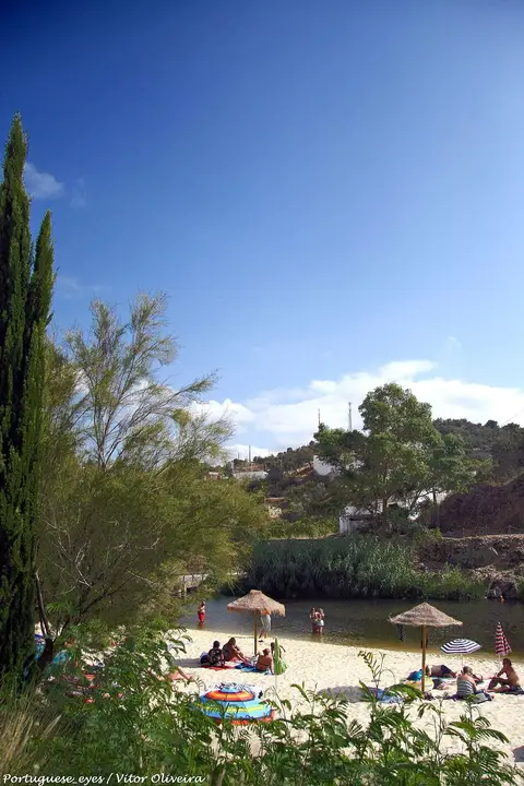 Praia fluvial do Pego Fundo, em Alcoutim, com areia, chapéus de palha e vegetação junto ao rio