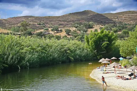 Praia fluvial do Pego Fundo, em Alcoutim, com areal, chapéus de palha e vegetação junto ao rio