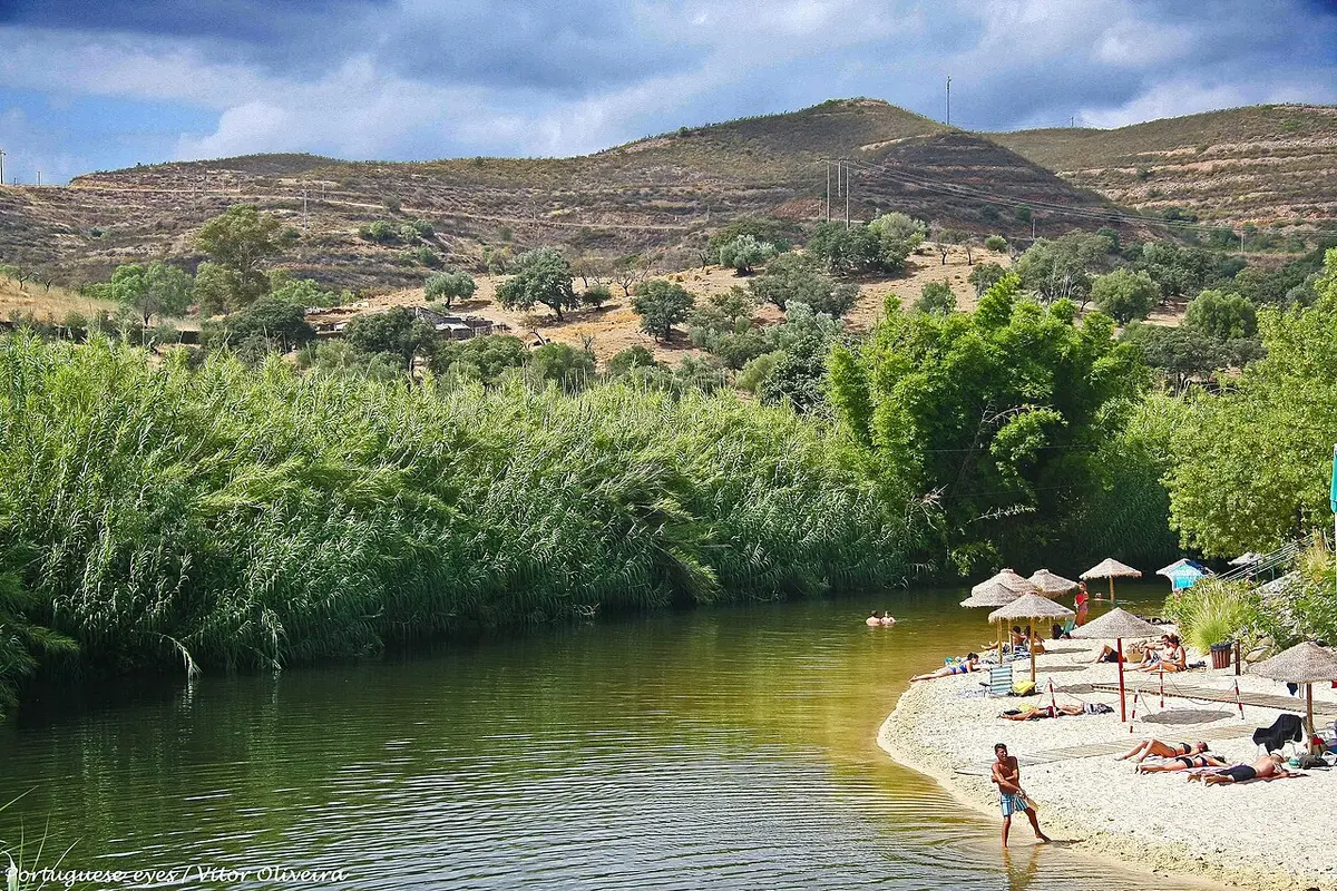 Praia fluvial do Pego Fundo, em Alcoutim, com areal, chapéus de palha e vegetação junto ao rio