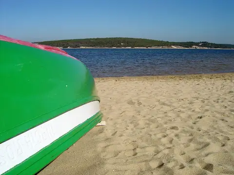 Barco verde virado na areia junto à água da Lagoa de Albufeira, com pinhal ao fundo