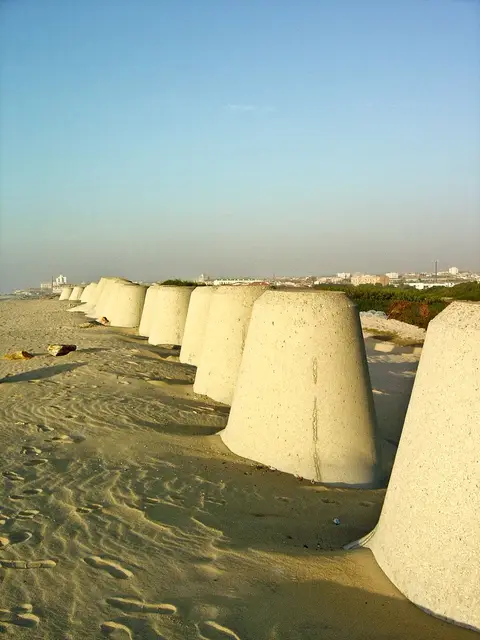Fila de blocos de betão cónicos sobre a areia da praia de Silvalde, com a cidade ao fundo
