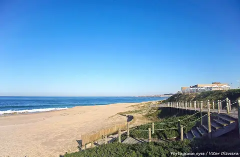 Praia do Aterro com areal extenso, mar calmo e passadiço de madeira com escadas entre a vegetação