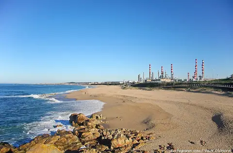 Praia do Aterro com areal extenso, rochas junto ao mar e chaminés da refinaria ao fundo