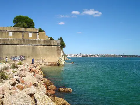 Zona rochosa junto ao muro do forte na praia de Albarquel, com água esverdeada e vista para a cidade