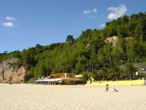 Praia de areia em Albarquel com restaurante amarelo junto à encosta arborizada e céu azul