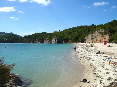 Praia de Albarquel com areia clara, água azul-turquesa e encosta arborizada com rochedos ao fundo