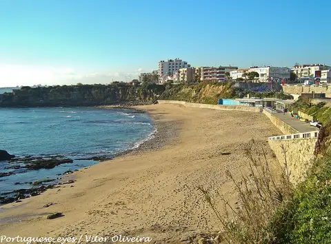 Praia de areia de São Pedro do Estoril ao lado de falésia e passeio marítimo, com edifícios ao fundo