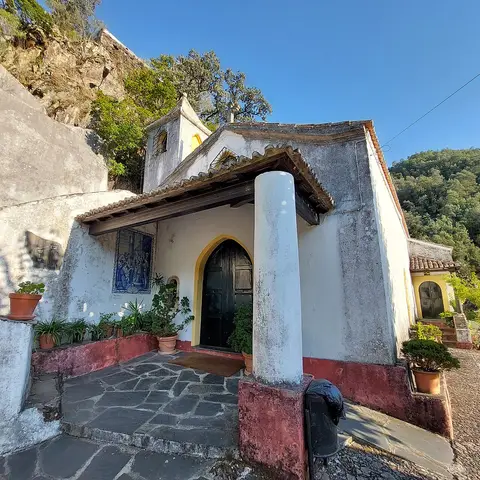 Capela branca com painel de azulejos e vasos de plantas, encostada à encosta rochosa da Lousã.
