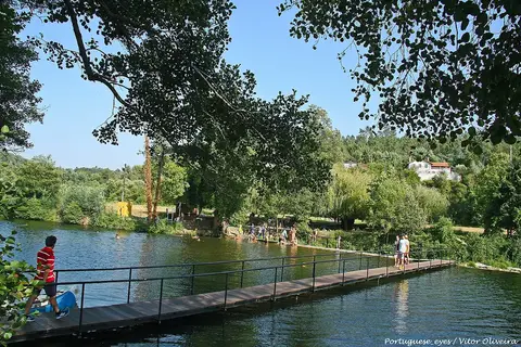 Praia fluvial da Bogueira com passadiço flutuante sobre o rio, rodeado de árvores.