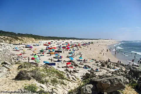 Praia de Pedrógão com areal extenso, chapéus de sol coloridos, rochas em primeiro plano e mar azul.