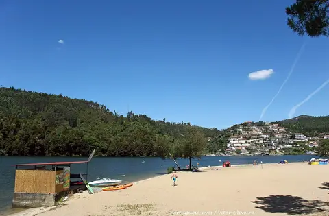 Praia fluvial de areia junto ao rio, com caiaques, barco e colinas arborizadas ao fundo