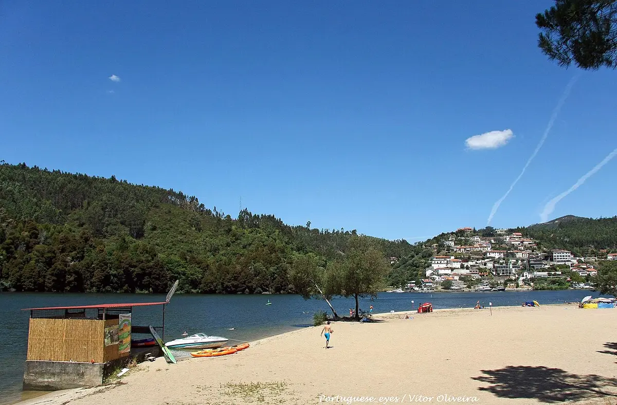 Praia fluvial de areia junto ao rio, com caiaques, barco e colinas arborizadas ao fundo