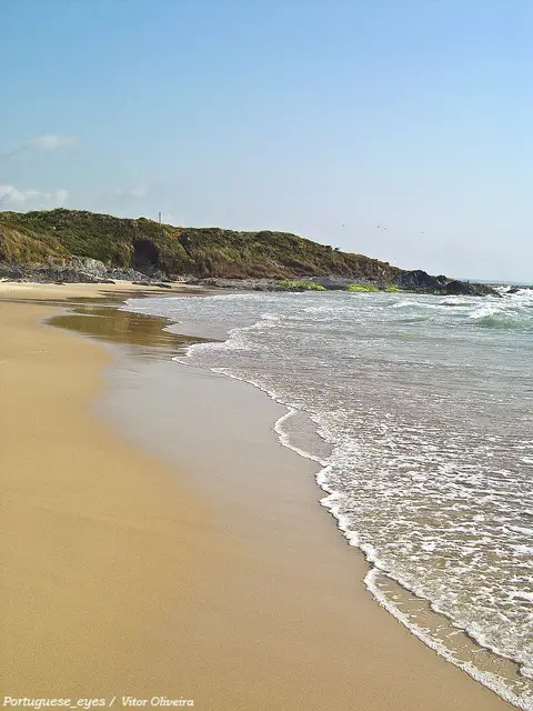 Praia de areia deserta com ondas suaves e promontório verde ao fundo, em Vale de Figueiros