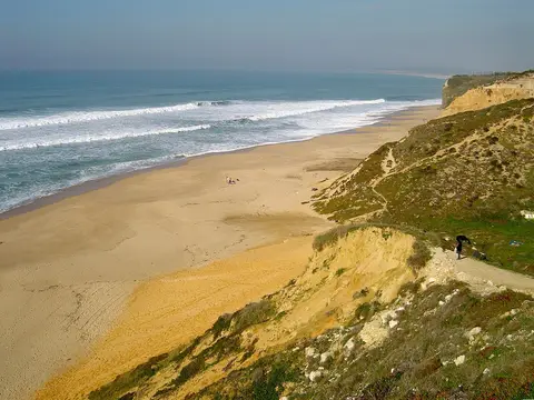 Vista do alto da arriba sobre a Praia das Bicas, com areal extenso e ondas a rebentar.