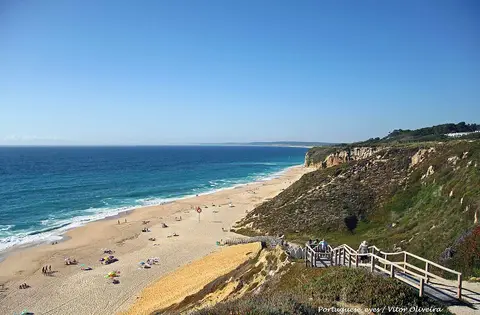 Vista da Praia das Bicas com areal extenso, falésias verdes e escadaria de acesso à praia.