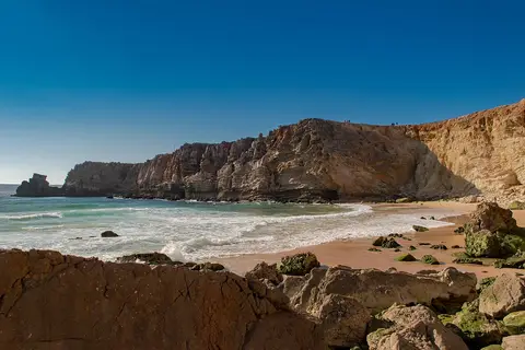Praia do Tonel com falésias douradas, rochas na areia e ondas a rebentar sob céu azul