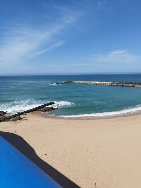 Praia de areia junto ao mar azul, com quebra-mar rochoso ao fundo, na Ericeira
