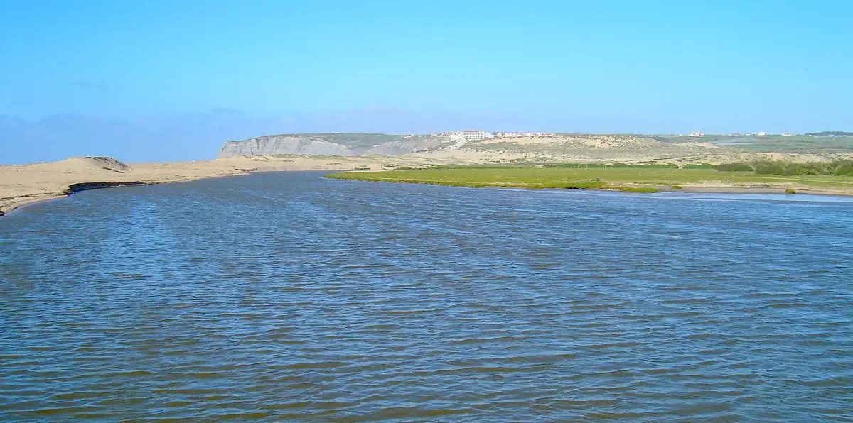 Foz do rio Sizandro, com areal, vegetação e falésias ao fundo sob céu azul