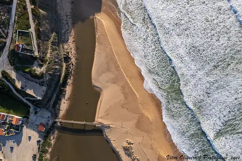 Vista aérea da foz do rio Sizandro, com praia de areia, passadiço e ondas do mar