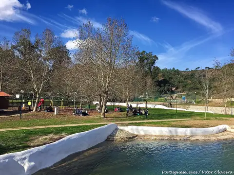 Praia fluvial dos Olhos de Água do Alviela, com relvado, árvores, parque infantil e margem caiada junto ao rio.