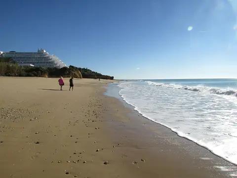 Praia da Oura com areal extenso, ondas suaves e edifícios brancos ao fundo num dia de céu limpo