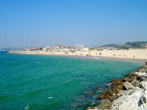 Praia da Cova do Vapor com areal, casas ao fundo, rochas do molhe e mar verde-azulado.