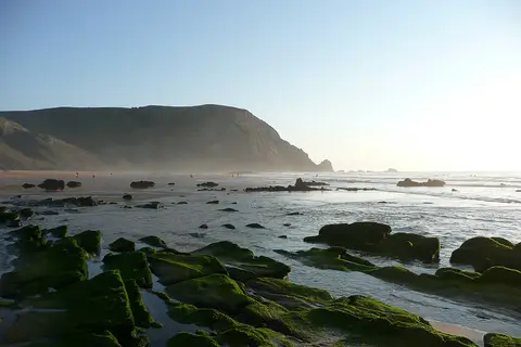 Praia do Castelejo com rochas cobertas de musgo verde na maré baixa e falésia ao fundo