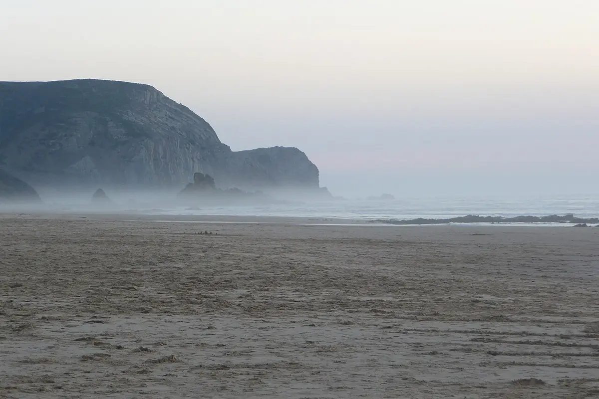 Praia do Castelejo ao amanhecer, com areia extensa, neblina e falésias sobre o mar.