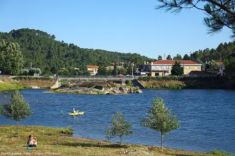 Praia fluvial em Vila da Ponte, com rio, ponte, árvores e casas ao fundo