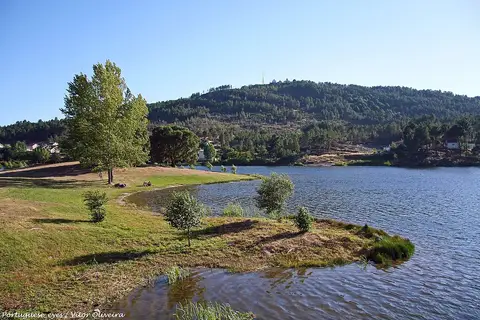 Albufeira da Barragem do Vilar com margem relvada, árvores e colinas arborizadas ao fundo