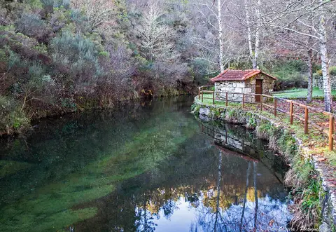 Rio de águas calmas e esverdeadas junto a pequena casa de pedra e vegetação, na praia fluvial de Pontemieiro