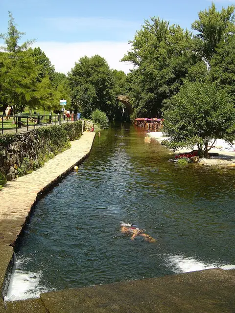 Praia fluvial com rio de águas claras, ponte de pedra em arco e passeio ladeado de árvores