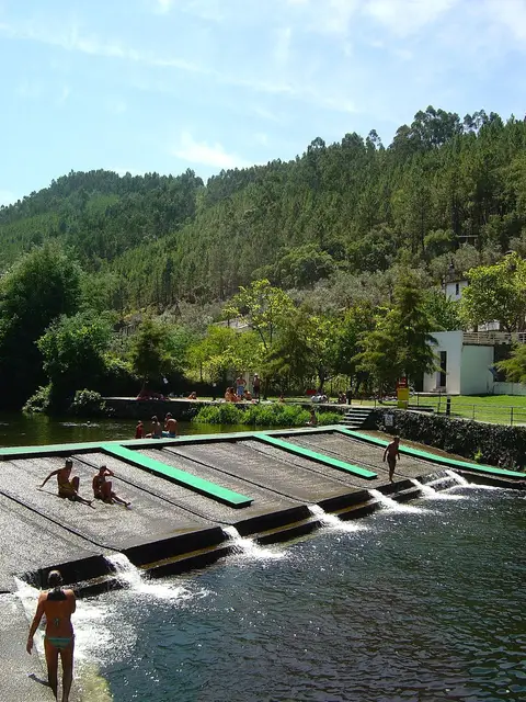 Praia fluvial com açude em socalcos e escorregas, rodeada de encostas arborizadas