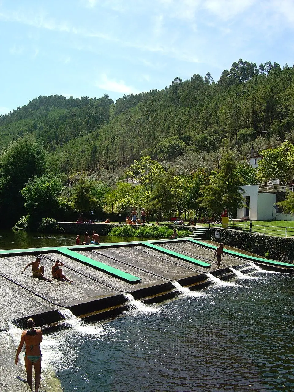 Praia fluvial com açude em socalcos e escorregas, rodeada de encostas arborizadas