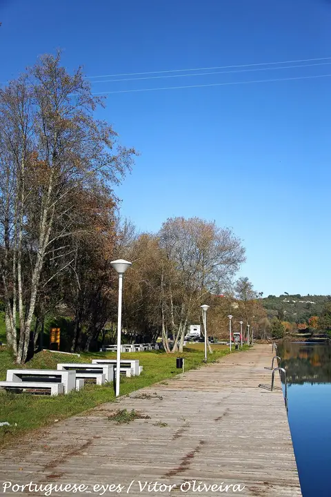 Deque de madeira junto ao rio na praia fluvial de Ponte de Juncais, com mesas de merendas e árvores.