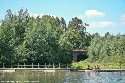 Praia fluvial das Canaveias com passadiço de madeira, rio calmo e ponte de pedra entre árvores