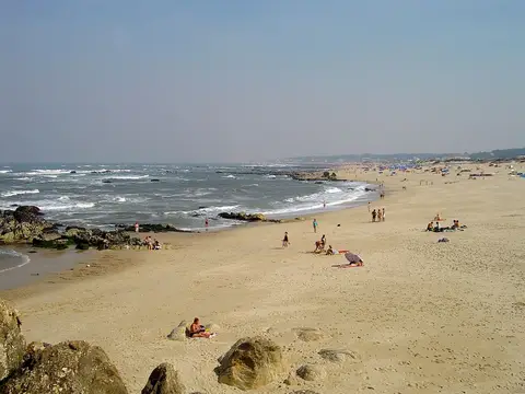 Praia de areia extensa com rochas e ondas do mar, com banhistas ao longe