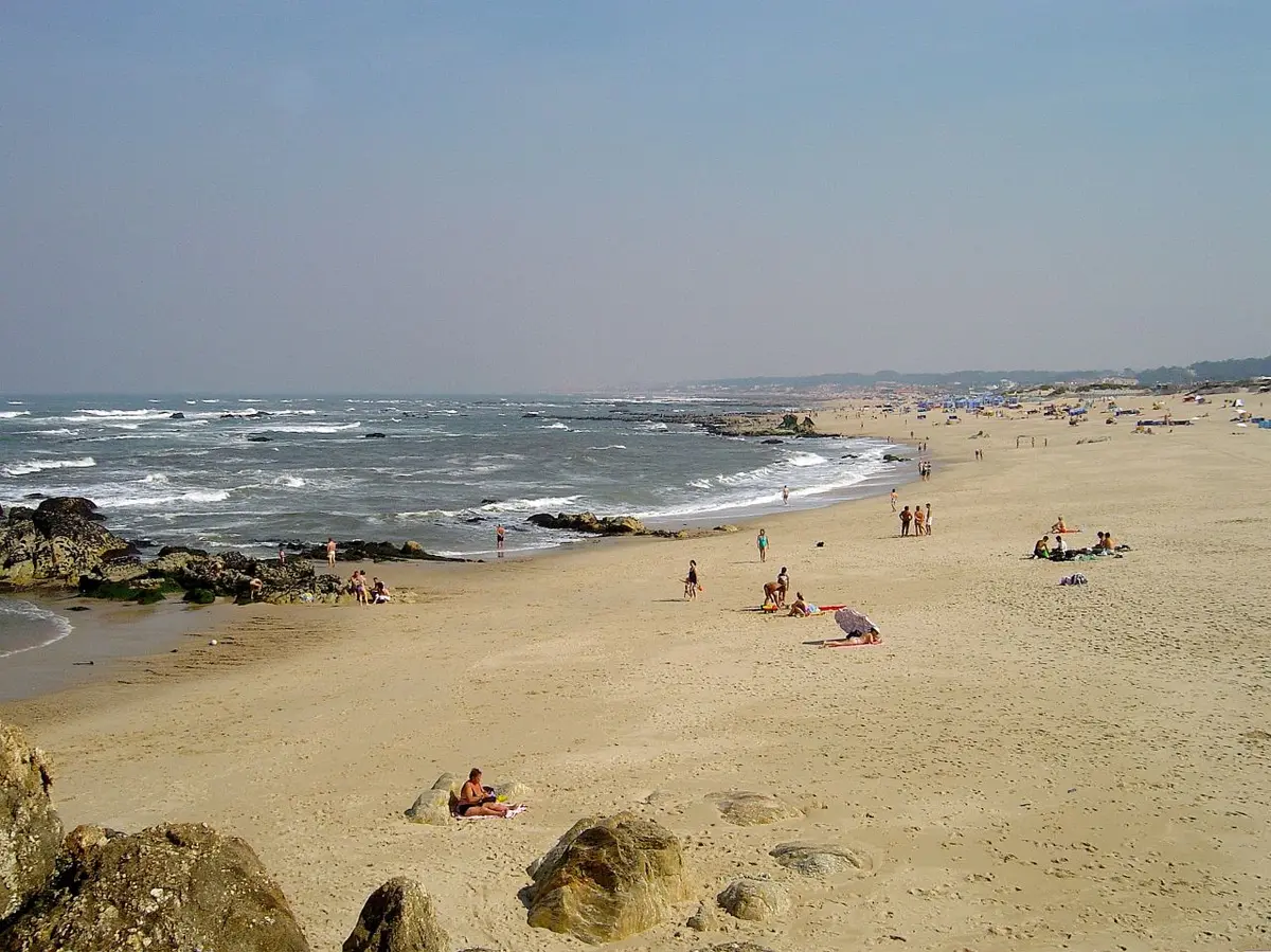 Praia de areia extensa com rochas e ondas do mar, com banhistas ao longe