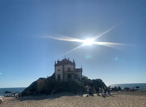 Capela do Senhor da Pedra sobre rochedo na praia, em contraluz com o sol ao alto