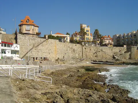 Praia da Azarujinha, no Estoril, com rochas, muralha de pedra e casas sobre a falésia.
