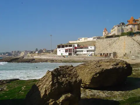 Costa rochosa junto à Praia da Azarujinha, com edifícios brancos e mar agitado ao fundo