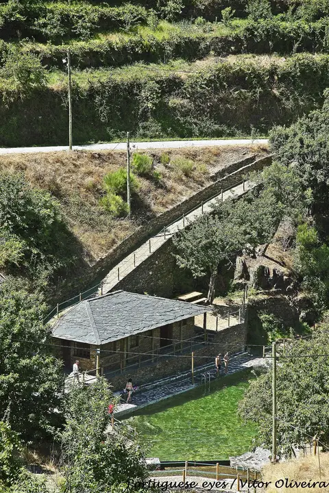 Praia fluvial junto a edifício de xisto, com piscina natural e caminho em encosta verde, em Piódão