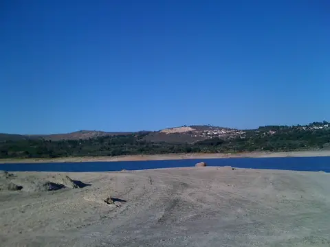 Margem arenosa da albufeira do Vilar, com água azul e colinas com aldeia ao fundo