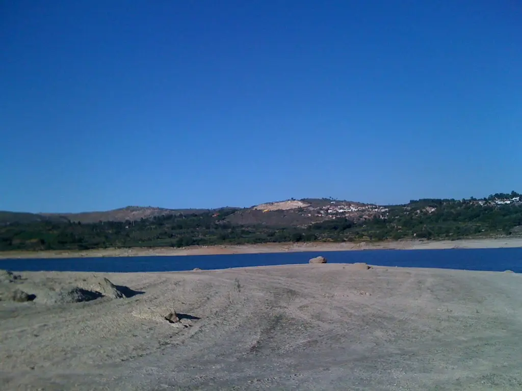 Margem arenosa da albufeira do Vilar, com água azul e colinas com aldeia ao fundo